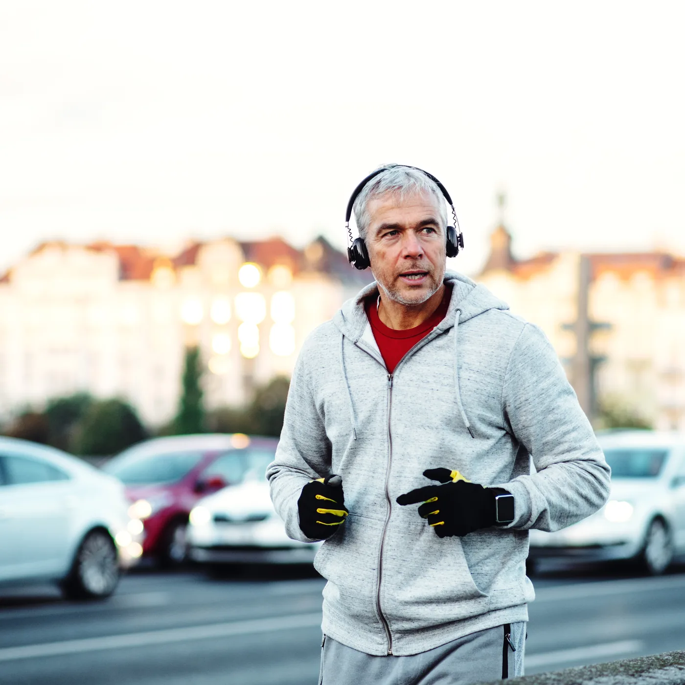 A grey-haired man in a hooded sweatshirt and headphones running through a city street